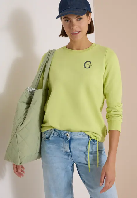 Frau trägt hellgrünen Pullover, Jeans, Cap und eine gesteppte Tasche.