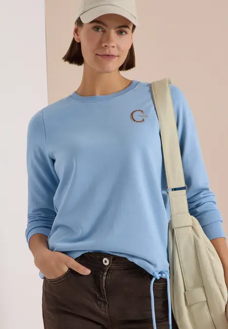 Eine Frau mit hellblauem Langarmshirt, dunkler Hose, hellbeiger Cap und Schultertasche.