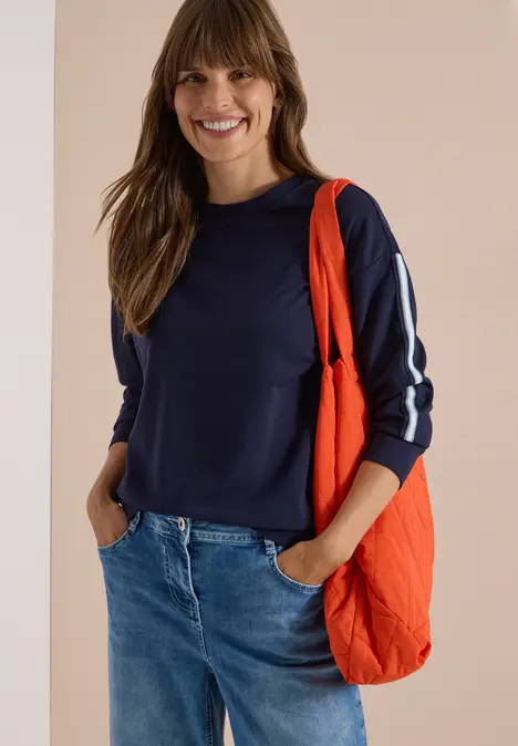 Frau in Jeans, dunkelblauem Sweatshirt mit weißen Streifen und oranger Tasche lächelt.