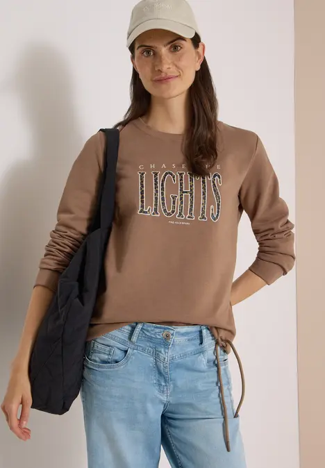 Junge Frau mit beigefarbener Cap, braunem Sweatshirt mit Schriftzug LIGHTS, Jeans und schwarzer Tasche.