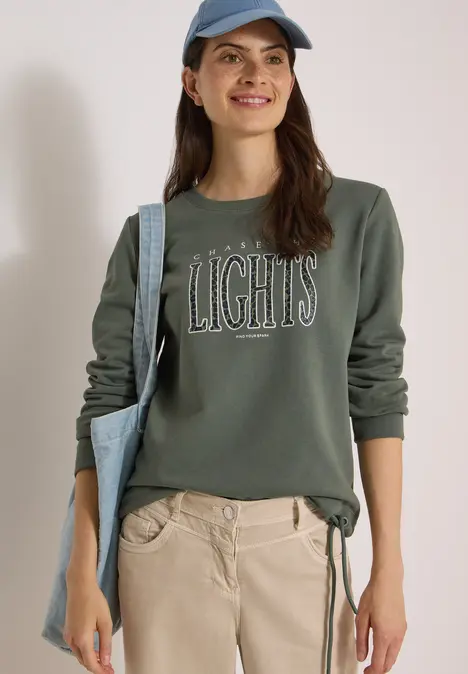 Frau lächelt, trägt ein olivgrünes Sweatshirt mit Aufschrift 'LIGHTS', helle Hose, blaue Cap und Tasche.