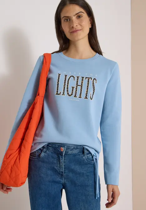 Frau trägt hellblauen Pullover mit 'LIGHTS'-Schriftzug, Jeans und orangefarbene Tasche vor hellem Hintergrund.