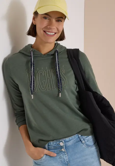 Eine lächelnde Frau trägt eine grüne Kapuzenjacke, gelbe Mütze und Jeans, sie trägt eine schwarze Tasche.