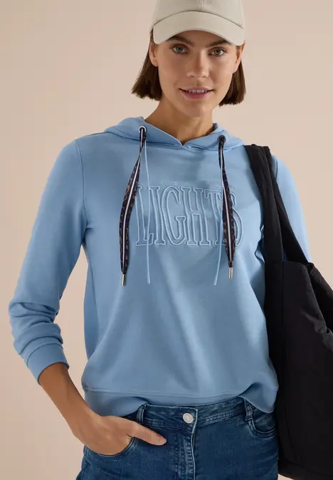 Frau in hellblauem Kapuzenpullover, Jeans, beiger Cap und schwarzer Tasche vor hellem Hintergrund.