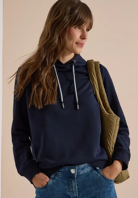 Eine Frau in dunkelblauer Kapuzenjacke, Jeans und Schultertasche, die seitlich in die Ferne blickt.