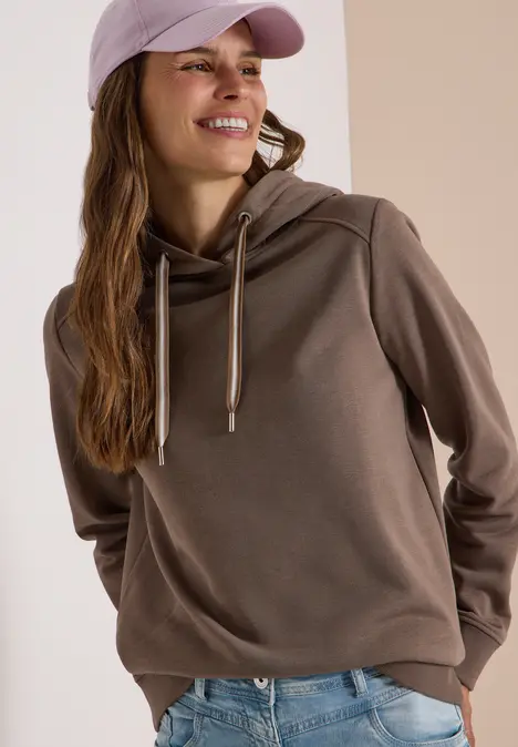 Junge Frau in Hoodie, Jeans und Baseballcap, lächelnd und seitlich blickend.