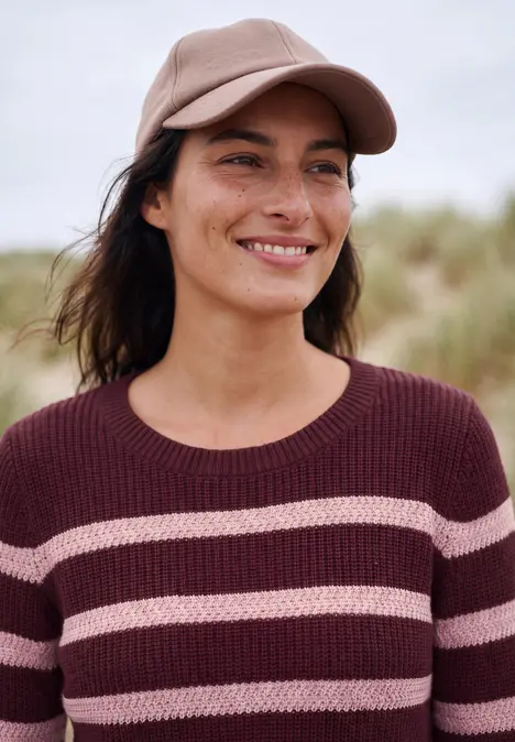 Lächelnde Frau mit Basecap und gestreiftem Pullover steht draußen vor verschwommenem Hintergrund.