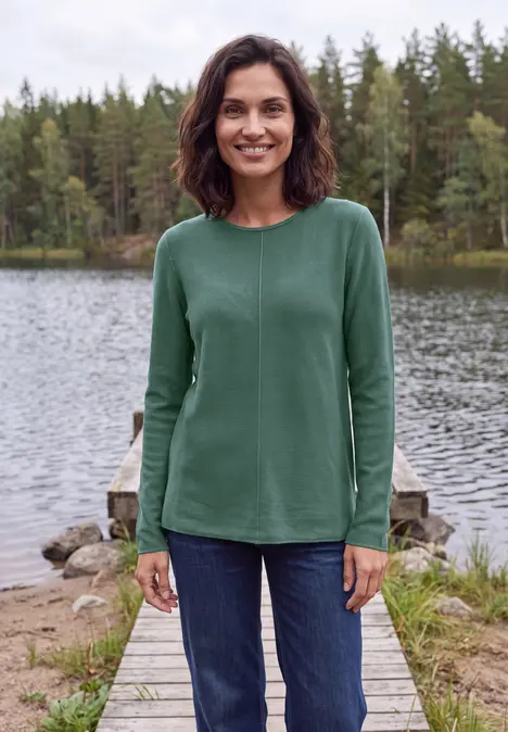Eine lächelnde Frau in grünem Pullover und Jeans steht auf einem Steg am See, umgeben von Wäldern.