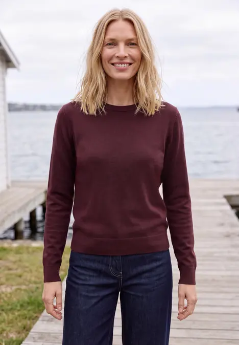 Eine lächelnde Frau in Pullover und Jeans steht auf einem Holzsteg am Wasser.