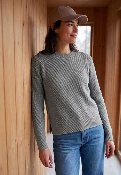 Eine Frau lehnt in grauem Pullover, blauer Jeans und Cap an einer Holzverkleidung und lächelt.