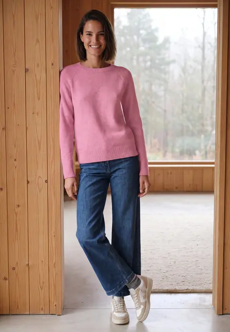 Eine lächelnde Frau in rosa Pullover, blauen Jeans und weißen Sneakern steht in einem hellen Holzraum.