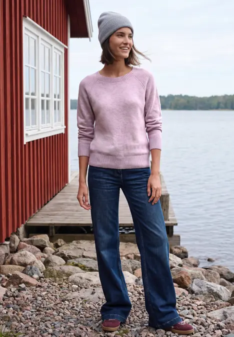 Frau in rosa Pullover und Mütze steht lächelnd in Jeans vor roter Hütte am See.