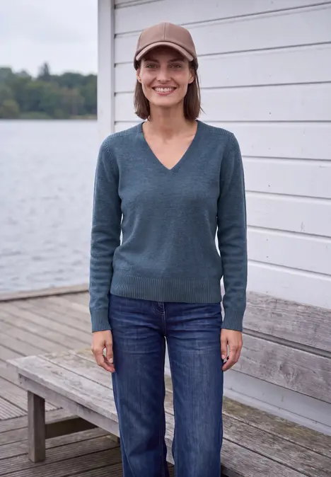 Eine Frau mit Mütze, Brille und blauem Pullover lächelt auf einem Steg am See vor einer weißen Holzwand.