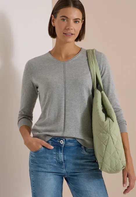 Eine Frau trägt einen grauen Strickpullover, blaue Jeans und eine olivgrüne Stepptasche über der Schulter.