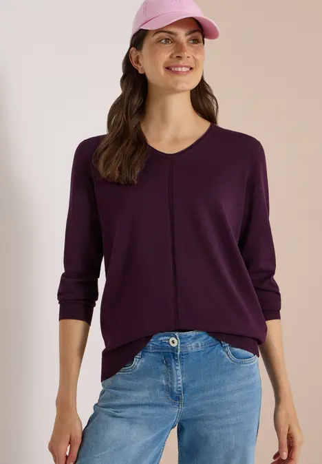 Eine lächelnde Frau in lila Pullover, Jeans und passender Cap vor neutralem Hintergrund.