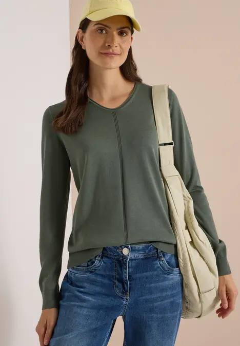 Eine junge Frau mit grünem Langarmshirt, Jeans, gelber Kappe und beiger Umhängetasche.
