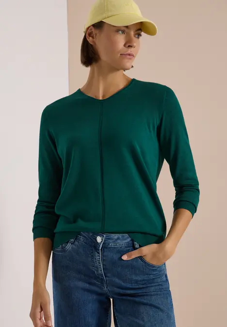Frau mit gelber Mütze und grünem Pullover, blaue Jeans, seitlich stehend vor hellem Hintergrund.