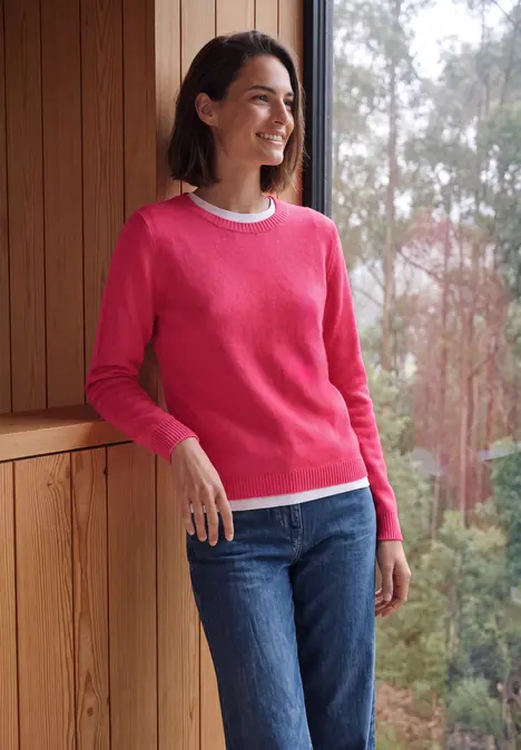 Frau in pinkem Pullover und Jeans steht lächelnd vor einem Holzrahmen mit Waldsicht.