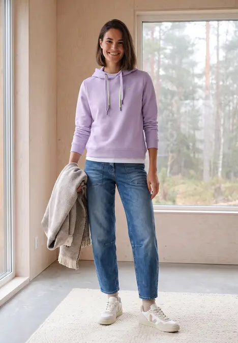 Frau in fliederfarbenem Hoodie, Jeans und Sneakern hält Jacke vor hellem Fenster.