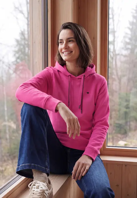 Frau in pinkem Kapuzenpullover und Jeans sitzt lächelnd am Fenster, Wald im Hintergrund.
