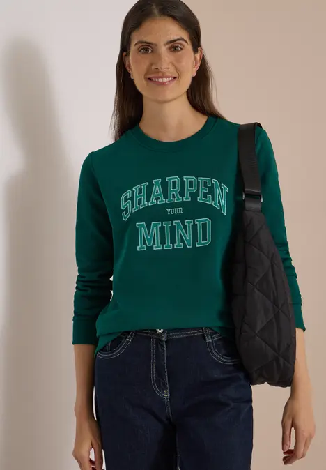 Uśmiechnięta kobieta ma na sobie ciemnozielony sweter z napisem 'SHARPEN MIND' i ciemne jeansy.