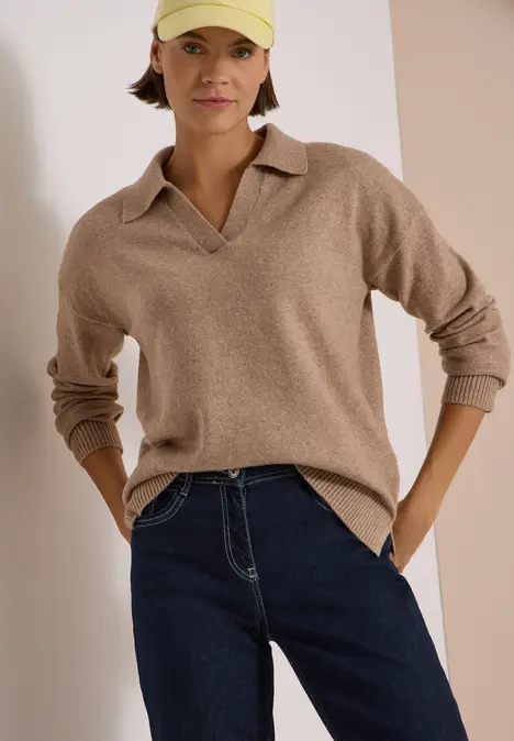 Frau trägt beigefarbenen Polokragen-Pullover, dunkle Jeans und gelbe Baseballkappe.