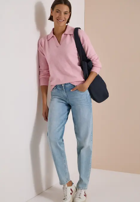 Eine lächelnde Frau lehnt an einer Wand und trägt rosa Pullover, helle Jeans und eine dunkelblaue Tasche.