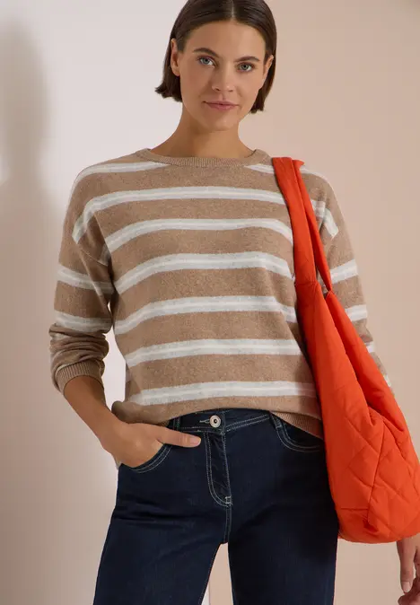 Frau in beige-weiß gestreiftem Pullover, Jeans und oranger Umhängetasche vor hellem Hintergrund.