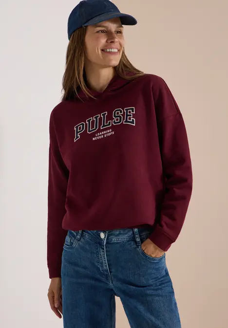 Lächelnde Frau mit burgunderroter Kapuzenjacke mit 'PULSE'-Schriftzug, Jeans und Baseballcap.