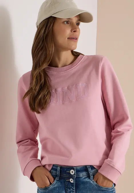 Frau trägt rosa Sweatshirt, blaue Jeans und beige Baseballkappe.