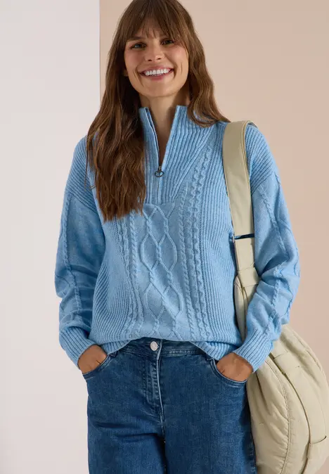 Lächelnde Frau in hellblauem Zopfpullover mit Reißverschluss, Jeans und beiger Tasche vor hellem Hintergrund.