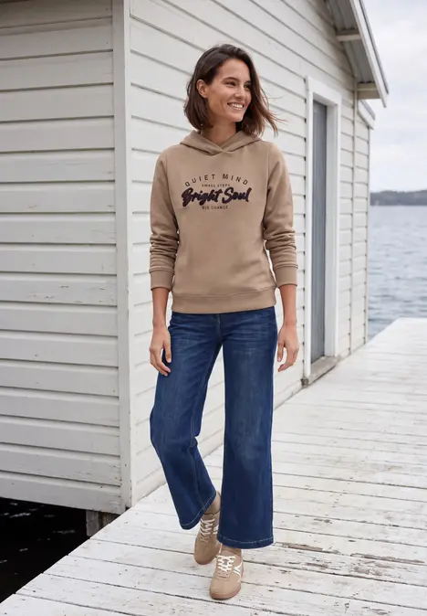 Frau in beigem Hoodie und Jeans spaziert auf Steg vor einem Haus am Wasser und lächelt.