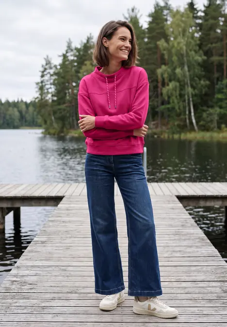 Eine Frau in pinkem Hoodie und Jeans steht lächelnd auf einem Steg am See mit Wald im Hintergrund.