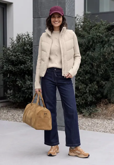 Frau mit lila Mütze, beiger Steppweste, Pullover, weiter Jeans, braunen Schuhen und Tasche vor Gebäude.