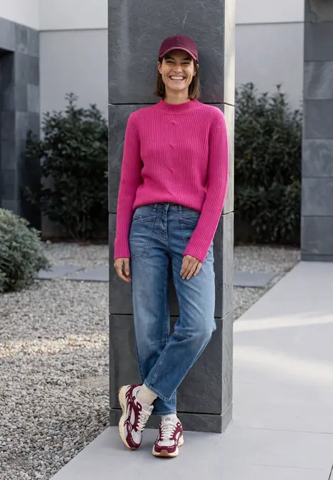 Eine lachende Frau in pinkem Pullover, Jeans und Cap lehnt vor einer Säule in modernem Außenbereich.