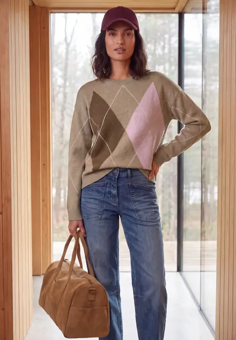 Frau mit roter Kappe, beigem Rautenpullover, blauen Jeans und brauner Tasche in hellem Raum vor Fenster.