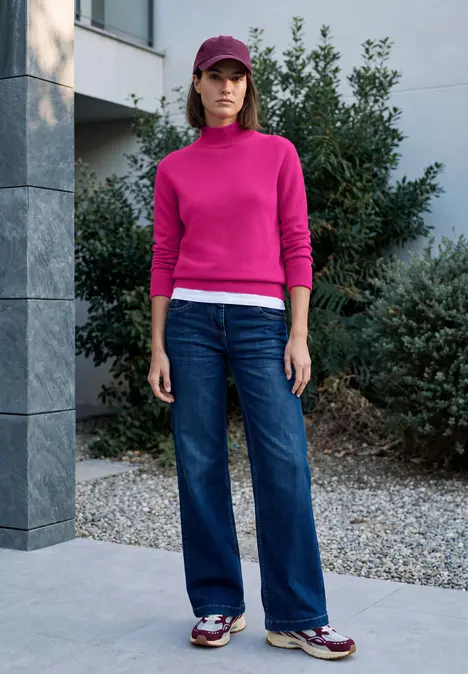 Frau in pinkem Pullover, Jeans, Sneakern und Cap steht vor modernem Haus mit Büschen.