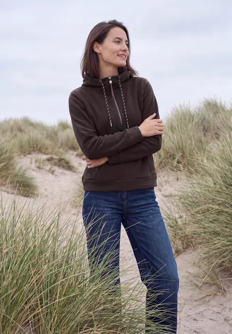 Eine Frau steht in Hoodie und Jeans mit verschränkten Armen in einer Dünenlandschaft am Strand.
