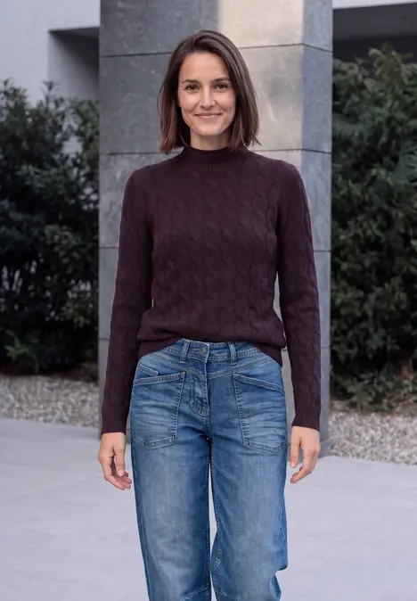 Eine Frau in dunkelrotem Strickpullover und Jeans steht vor einem Gebäude und lächelt leicht.