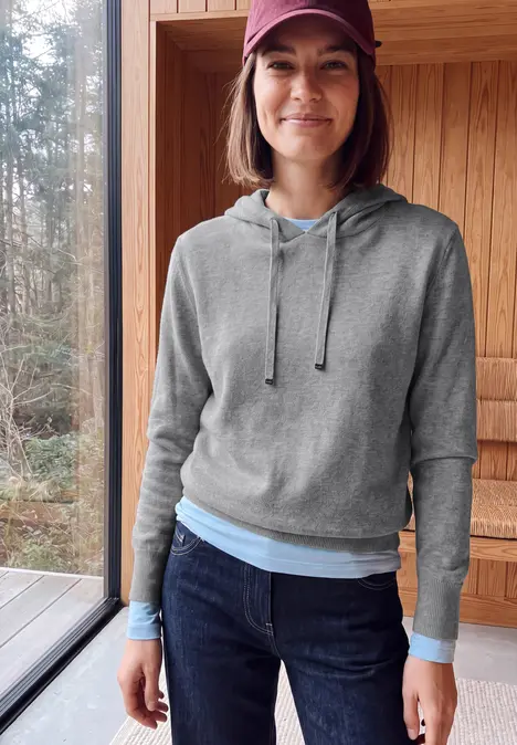 Frau in grauer Kapuzenjacke, Jeans und roter Mütze steht lächelnd in einer Holzhütte mit Waldblick.