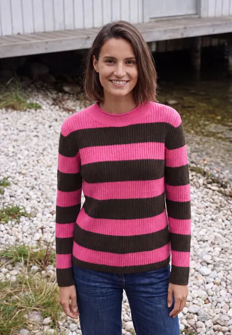 Frau mit rosa-grün gestreiftem Pullover und Jeans steht lächelnd am Ufer vor einem Holzsteg.