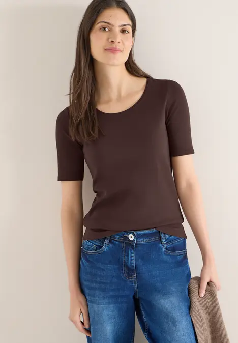 Frau trägt braunes T-Shirt und blaue Jeans, steht locker mit einem beigen Schal in der Hand.