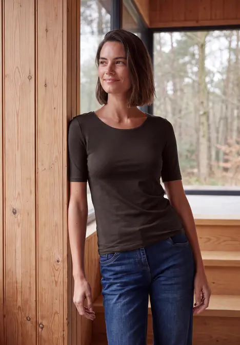 Kobieta opiera się o drewnianą ścianę, z widokiem na las, ma na sobie dżinsy i ciemny t-shirt.