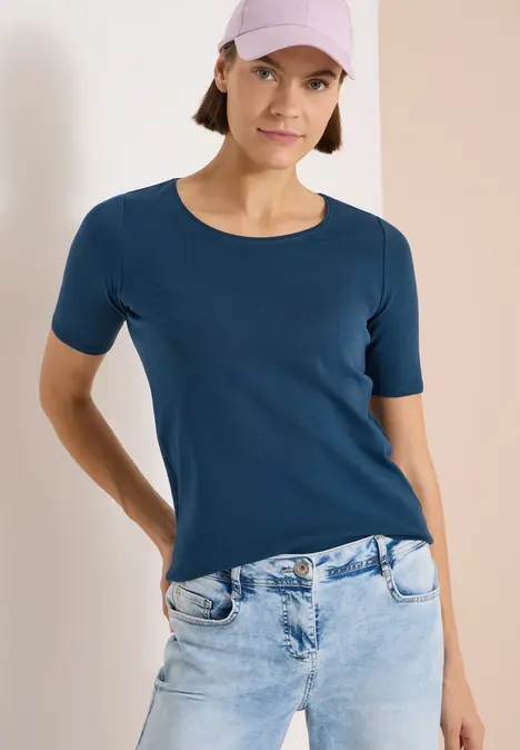 Frau trägt dunkelblaues T-Shirt und hellblaue Jeans, frontal abgebildet, Hände an den Hüften.