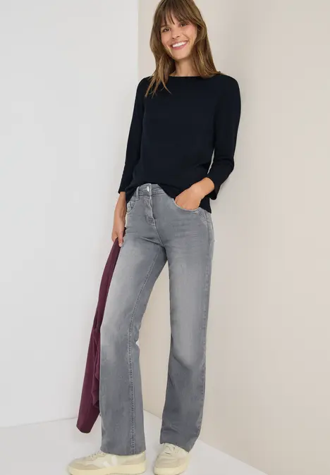 Frau in schwarzem Pullover, grauen Schlagjeans und weißen Sneakern, hält eine fliederfarbene Jacke.