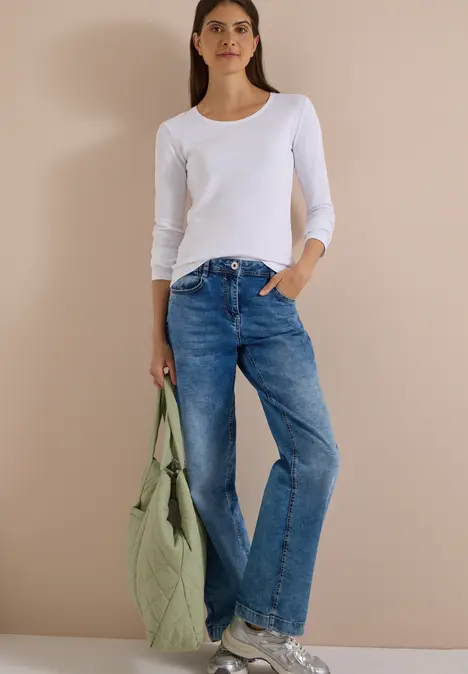 Frau trägt weißes Langarmshirt, blaue Jeans und hält eine gesteppte grüne Tasche.