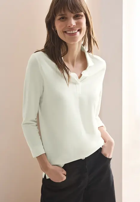 Frau mit hellem Langarmshirt und dunkler Hose lächelt, Hände in den Taschen.