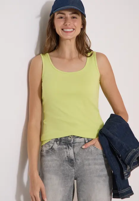 Eine Frau trägt ein gelbes Top, graue Jeans und hält eine dunkelblaue Jacke in der Hand.