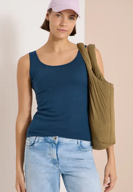 Frau in blauem Tanktop und hellblauen Jeans mit olivgrünem Schulterbeutel vor neutralem Hintergrund.