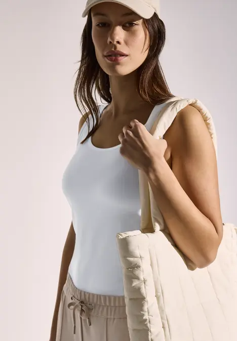 Frau mit weißem Top, beigener Kappe und großer Tasche schaut in die Kamera.